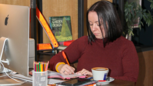 Gemma at desk writing in a notepad with a coffee next to her and a pot of colourful pencils